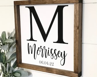 May include: A framed wooden sign with a white background. The sign features a large black letter "M" at the top, the name "Morrissey" in cursive below, and the date "06.04.22" in a smaller font. The frame is a dark brown color.