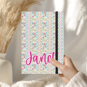 May include: A personalized notebook with the name "Janet" in pink script. The cover features a colorful pattern of pink, blue, and yellow designs. A black elastic band secures the notebook.
