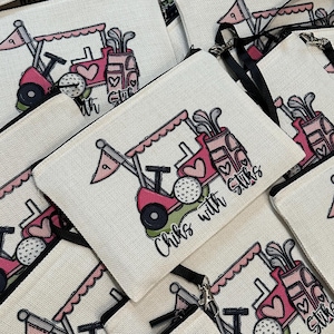 May include: White canvas zippered pouch with a pink golf cart design, golf balls, and the text "Chiks with Stiks." The design includes hearts and golf clubs. The pouch has a black zipper and a black wrist strap.