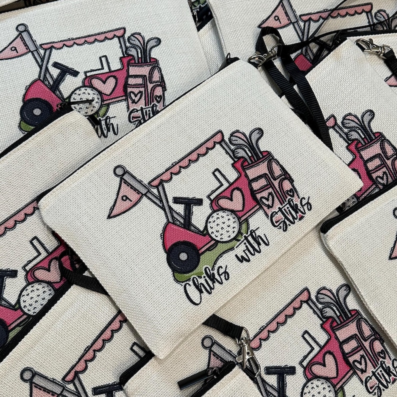 May include: White canvas zippered pouch with a pink golf cart design, golf balls, and the text "Chiks with Stiks." The design includes hearts and golf clubs. The pouch has a black zipper and a black wrist strap.