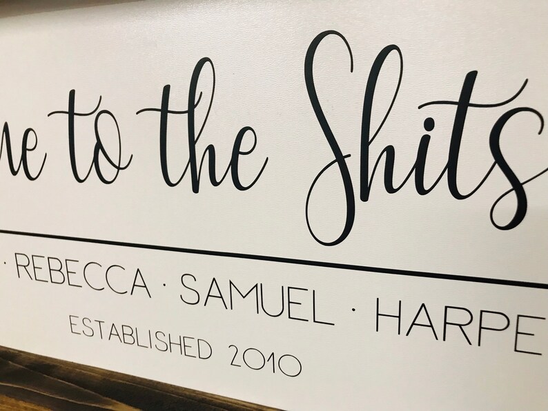 May include: White wooden sign with black calligraphy that reads "Welcome to the Shits - Rebecca - Samuel - Harpe Established 2010".