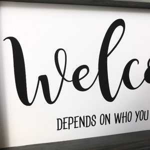 May include: A white canvas sign with a black cursive welcome sign and the text "depends on who you are asking" underneath.