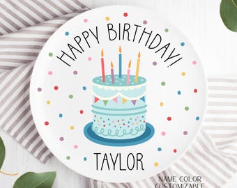 Puede incluir: Plato de cerámica blanco con un pastel de cumpleaños azul y blanco con velas y confeti de colores. El texto "Happy Birthday!" está escrito en negro alrededor del pastel y el nombre "Taylor" está escrito debajo del pastel.