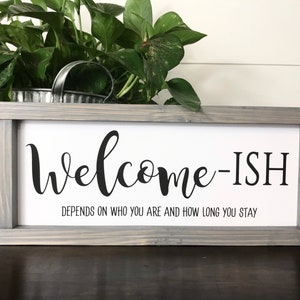 May include: A gray wooden framed sign with the text "Welcome-ish" and the caption "Depends on who you are and how long you stay".