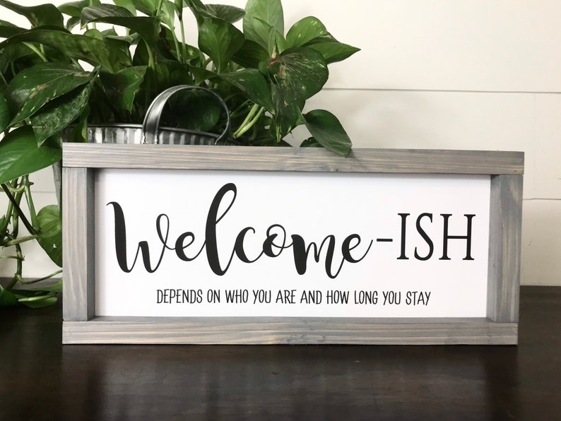 May include: A gray wooden framed sign with the text "Welcome-ish" and the caption "Depends on who you are and how long you stay".