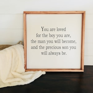 May include: A wooden sign with a white background and black text that reads "You are loved for the boy you are, the man you will become, and the precious son you will always be."