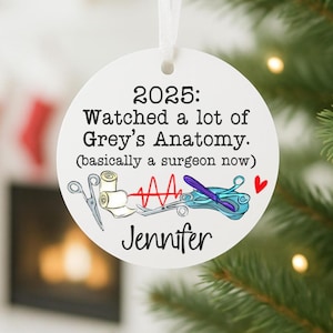 May include: A white, round Christmas ornament with the text "2025: Watched a lot of Grey's Anatomy. (basically a surgeon now)". The ornament features medical-themed illustrations and the name "Jennifer".