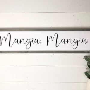 May include: A white wooden sign with a gray frame, featuring the text "Mangia, Mangia" in black cursive lettering.