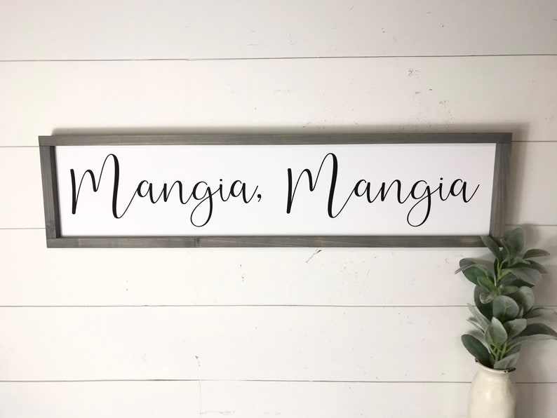 May include: A white wooden sign with a gray frame, featuring the text "Mangia, Mangia" in black cursive lettering.