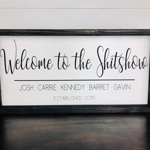 May include: A white wooden sign with black text that reads "Welcome to the Shitshow" with the names Josh, Carrie, Kennedy, Barret, and Gavin listed below and the date "Established 2019".