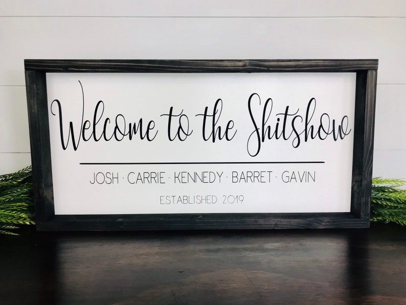 May include: A white wooden sign with black text that reads "Welcome to the Shitshow" with the names Josh, Carrie, Kennedy, Barret, and Gavin listed below and the date "Established 2019".