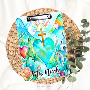 May include: A clipboard with a colorful watercolor design featuring a cross, heart, church, and rainbow. The clipboard has a metal clip at the top and the name "Ms. Nicole" is written on the bottom. The background is a woven basket.