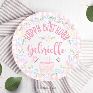 May include: A pink and white cake plate with a gingham pattern and floral border. The plate says "Happy Birthday Gabrielle" in pink script and features a pink cake with candles and butterflies.