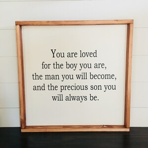 May include: A framed sign with a white background and a wooden frame. The sign reads "You are loved for the boy you are, the man you will become, and the precious son you will always be."