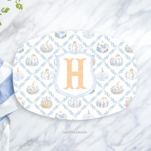 May include: Oval serving platter with a white background, featuring a repeating pattern of blue floral pumpkins and a light blue trellis design. A central shield displays a large, orange letter "H". The text "carolinamade" is at the bottom.