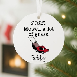 May include: A white, round Christmas ornament with the text "2025: Mowed a lot of grass." and a red and black lawnmower graphic. The name "Bobby" is written below the text. A white ribbon is attached for hanging.