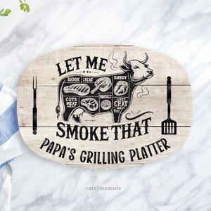 May include: A white oval wooden serving platter with a black and white illustration of a cow with different cuts of meat labeled. The text "Let Me Smoke That Papa's Grilling Platter" is printed on the platter.