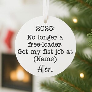 May include: A white, round Christmas ornament with the text "2025: No longer a free-loader. Got my fist job at (Name) Allen" in black. The ornament hangs from a white ribbon, set against a blurred Christmas tree background.