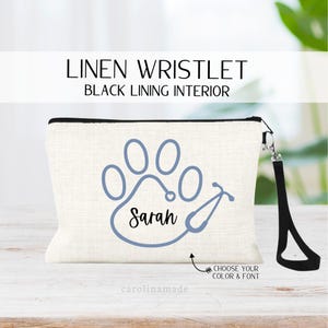 May include: A white linen wristlet with a black lining. The wristlet features a blue paw print with a stethoscope wrapped around it and the name "Sarah" written inside the paw print. The wristlet has a black zipper and a black wrist strap.