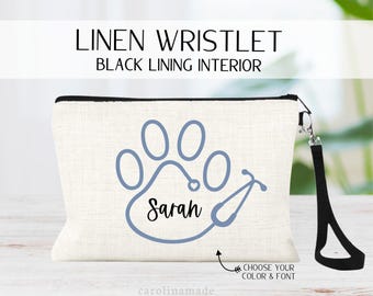May include: A white linen wristlet with a black lining. The wristlet features a blue paw print with a stethoscope wrapped around it and the name "Sarah" written inside the paw print. The wristlet has a black zipper and a black wrist strap.