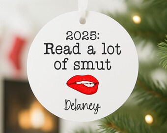 May include: A white, round Christmas ornament with the text "2025: Read a lot of smut" in black. A red lip graphic is below the text, and the name "Delaney" is written below the lip graphic. A white ribbon is attached for hanging.