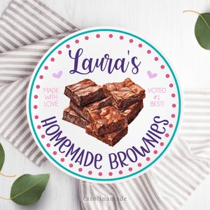 May include: A white round plate with a teal border and pink polka dots. The plate has the text "Laura's Homemade Brownies" in purple lettering. The text "Made with Love" and "Voted #1 Best" are also on the plate. A pile of chocolate brownies is in the center of the plate.