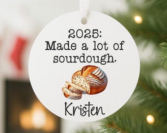 May include: White round Christmas ornament with the text "2025: Made a lot of sourdough." and the name "Kristen." The ornament features a watercolor illustration of a loaf of bread and sliced bread.