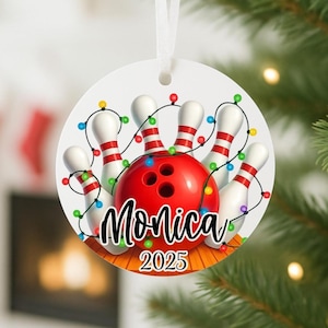 May include: A round Christmas ornament featuring a bowling theme. The design includes a red bowling ball, white bowling pins with red stripes, and colorful string lights. The name "Monica" and the year "2025" are also displayed.