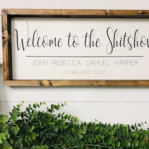 May include: A wooden sign with a white background and a brown frame. The sign says "Welcome to the Shitshow" with the names "John, Rebecca, Samuel, Harper" and "Established 2010" underneath.