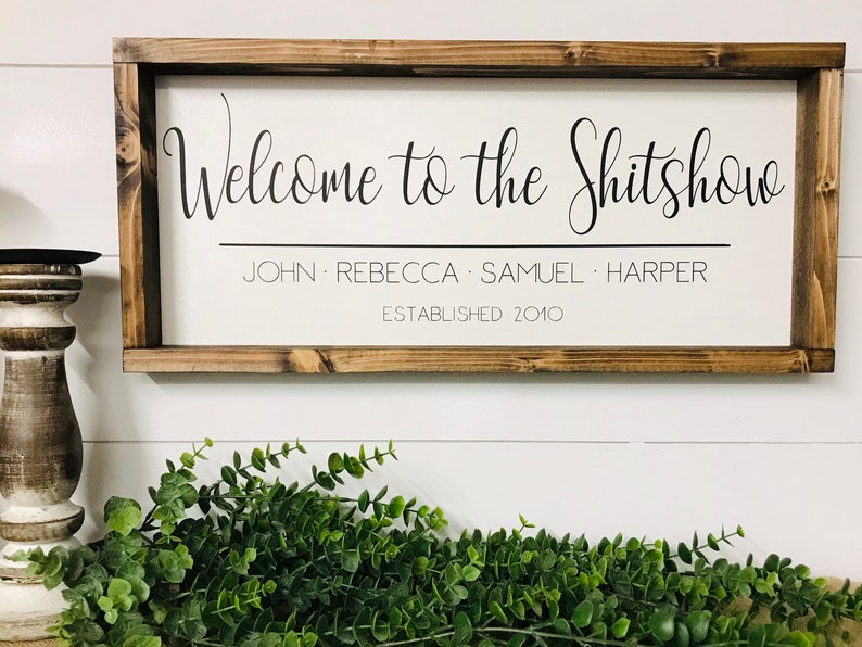 May include: A wooden sign with a white background and a brown frame. The sign says "Welcome to the Shitshow" with the names "John, Rebecca, Samuel, Harper" and "Established 2010" underneath.