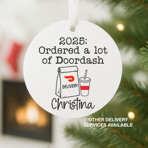 May include: White round Christmas ornament with the text "2025: Ordered a lot of Doordash" and a graphic of a delivery bag and drink. The name "Christina" is printed below the graphic. A white ribbon is attached for hanging.