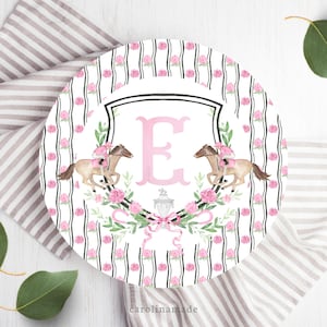 May include: A white round plate with a black and white striped border. The center of the plate features a pink letter "E" in a black and white shield with two pink horses and riders. The shield is surrounded by a wreath of pink flowers and green leaves.