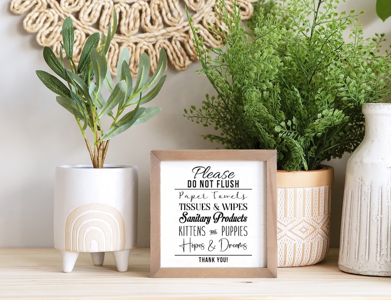 May include: A wooden sign with the text "Please Do Not Flush Paper Towels, Tissues & Wipes, Sanitary Products, Kittens & Puppies, Hopes & Dreams, Thank You!" in black lettering on a white background. The sign is sitting on a wooden surface with two potted plants.