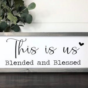 May include: A rectangular wooden sign with a gray frame. The white interior features the text "This is us" in a cursive font, with a small black heart. Below, the words "Blended and Blessed" are printed in a block font.