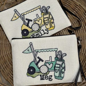 May include: Two canvas zipper pouches with golf-themed designs. One is yellow and the other is teal, both featuring golf carts, golf balls, and golf club bags. The yellow pouch has the word "family" and the teal pouch has the word "Meg".