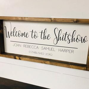 May include: A wooden sign with a white background and black text that reads "Welcome to the Shitshow John Rebecca Samuel Harper Established 2010".