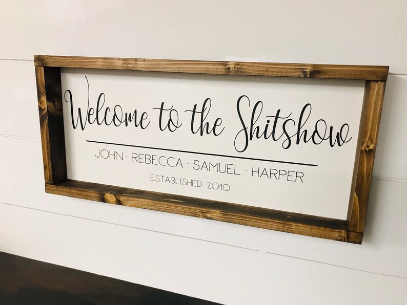 May include: A wooden sign with a white background and black text that reads "Welcome to the Shitshow John Rebecca Samuel Harper Established 2010".