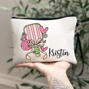 May include: A white canvas makeup bag featuring a pink and green paddle and a pink and white polka dot ball. The bag has a black zipper and a black strap. The name "Kristin" is written in black cursive on the bag.