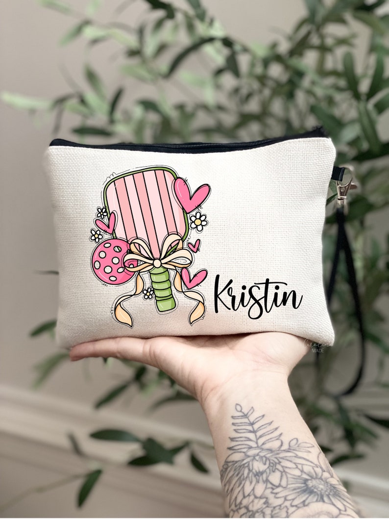 May include: A white canvas makeup bag featuring a pink and green paddle and a pink and white polka dot ball. The bag has a black zipper and a black strap. The name "Kristin" is written in black cursive on the bag.