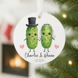 May include: A round, white ornament featuring two anthropomorphic pickles, one in a top hat and the other in a veil. The ornament has the names "Charlie & Renee" and the year "-2025-" printed on it, with pink hearts on either side. A white ribbon is attached for hanging.