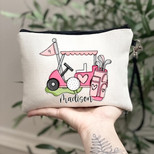 May include: A beige canvas zippered pouch featuring a pink golf cart design with a golf ball, golf clubs, and the name "Madison" in script. The golf cart has a heart-shaped detail. The pouch has a black zipper and a black wrist strap.