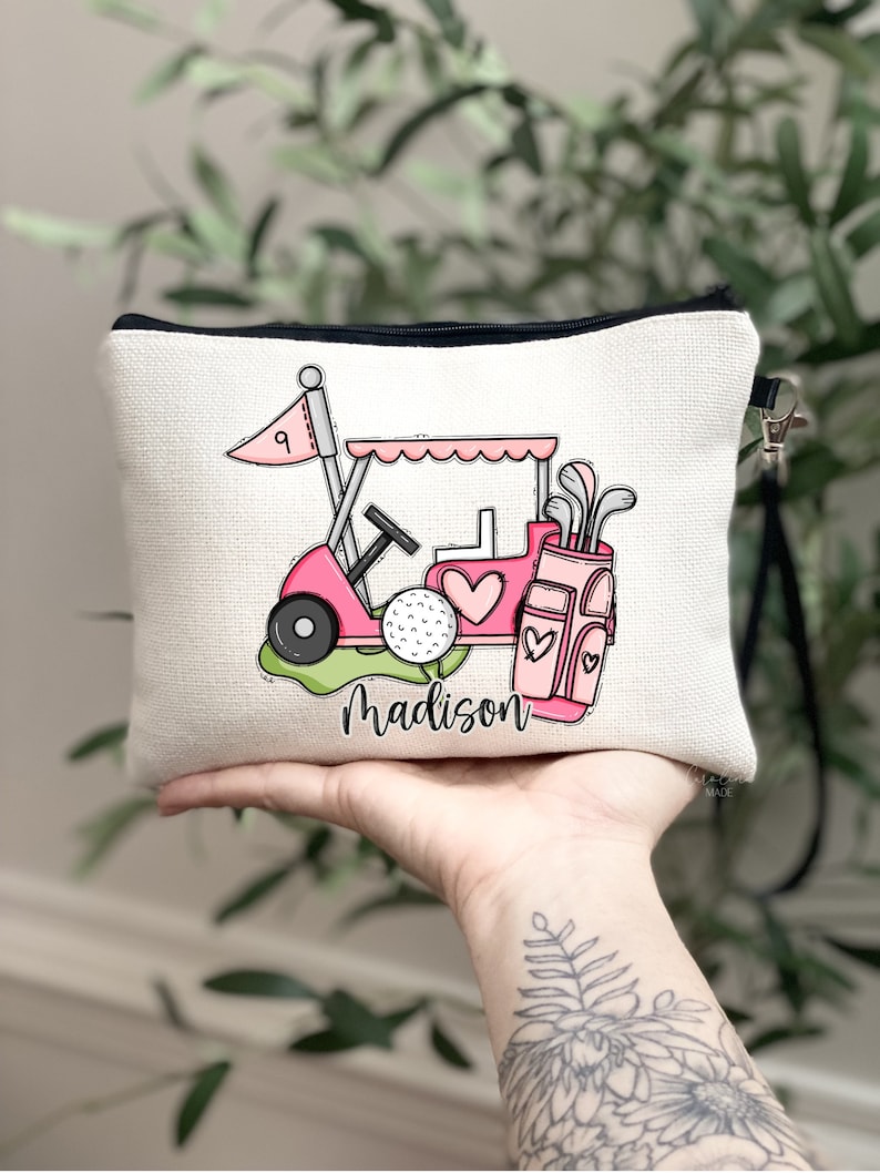 May include: A beige canvas zippered pouch featuring a pink golf cart design with a golf ball, golf clubs, and the name "Madison" in script. The golf cart has a heart-shaped detail. The pouch has a black zipper and a black wrist strap.