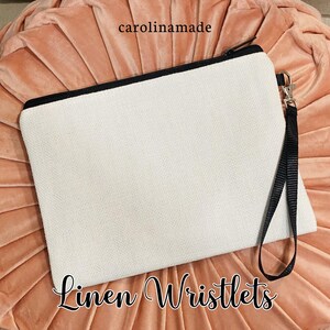May include: A white linen wristlet with a black zipper and a black strap. The wristlet is on a pink background. The text "Linen Wristlets" is written below the wristlet.