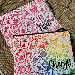 Puede incluir: Dos estuches de cremallera de lona blanca con un patr&oacute;n de fichas de mahjong de colores. El estuche superior tiene el nombre "Lisa" bordado. El estuche inferior tiene el nombre "Cheryl" bordado.