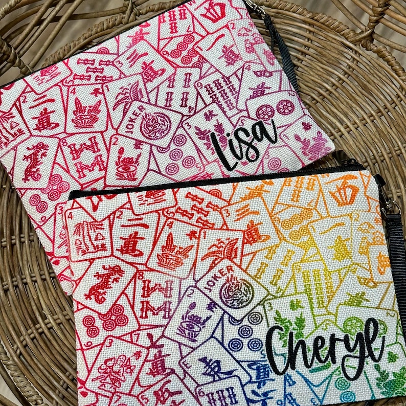 Puede incluir: Dos estuches de cremallera de lona blanca con un patr&oacute;n de fichas de mahjong de colores. El estuche superior tiene el nombre "Lisa" bordado. El estuche inferior tiene el nombre "Cheryl" bordado.