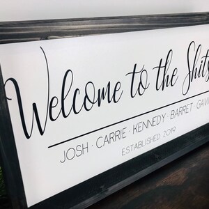 May include: A white sign with black text that reads "Welcome to the Shitshow Josh, Carrie, Kennedy, Barret, Gavin Established 2019". The sign is framed in a black wood frame.