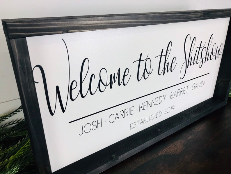 May include: A white sign with black text that reads "Welcome to the Shitshow Josh, Carrie, Kennedy, Barret, Gavin Established 2019". The sign is framed in a black wood frame.