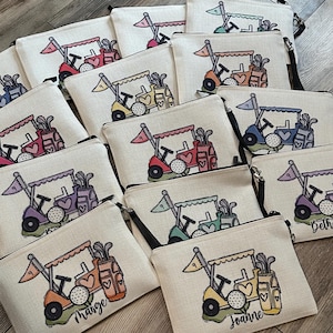 May include: Beige zippered pouches with a golf cart design in various colors. Each pouch features a golf cart with a flag, golf clubs, and a golf ball. Some pouches have names like "Marge" and "Joanne" printed on them.