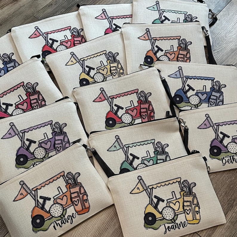 May include: Beige zippered pouches with a golf cart design in various colors. Each pouch features a golf cart with a flag, golf clubs, and a golf ball. Some pouches have names like "Marge" and "Joanne" printed on them.