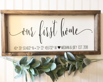 May include: A white wooden sign with the text "our first home" in black cursive script. Below the text are the coordinates "40° 39' 50.4612" N 73° 22′ 49.5732" W" and the text "MEGHAN & JOEY EST. 2018".